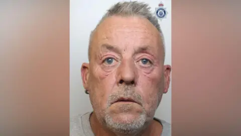 The mugshot of Gavin Shaw, who is wearing a grey t-shirt. He is starting forward and has short grey hair and a goatee. 