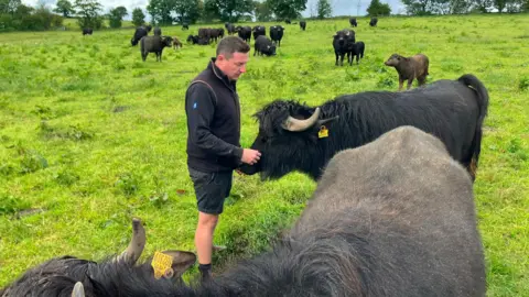 A man stands in a green field surrounded by water buffalos. He is wearing a long-sleeved black top with a black gilet over the top and black shorts.
