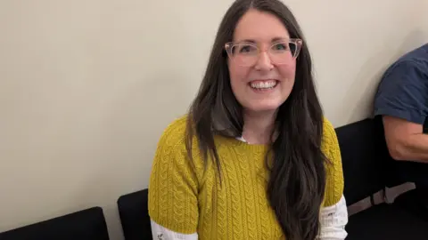 LDRS A woman wearing a yellow jumper with white sleeves sits on a chair, whilst smiling into the camera.