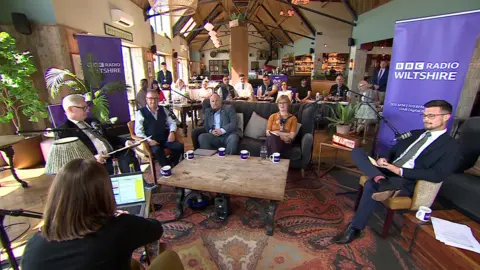 Five people sitting in front of microphones with purple signs which say 'BBC Radio Wiltshire' on them. They also have branded pull-up banners behind them and branded mugs on the coffee table in front of them. There is a Persian style rug on the floor and they are sitting in what looks like a large cafe.