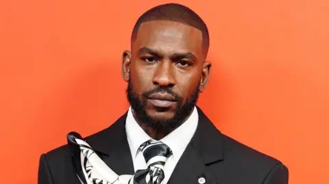 Skepta at the GQ Men of the Year awards. He is wearing a black suit and stood against a bright orange background.