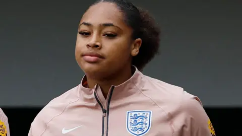 England goalkeeper Khiara Keating, with black hair scraped back from her face and wearing a pink tracksuit top bearing the England badge on it