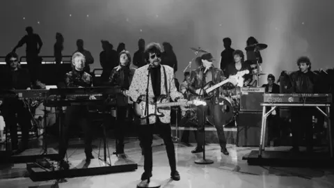 Tandy is on stage with other band members in this black and white photo. He is sat at a keyboard while other band members sing and play instruments.
