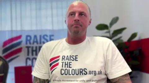 A man wearing a white t-shirt with text referring to Raise The Colours sits in front of a camera.