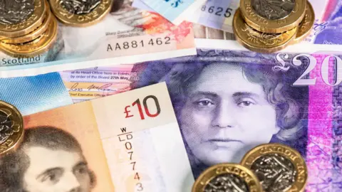 A collection of banknotes of different denominations, including £10 and £20. The colours of the notes include brown, pink and purple. On top of the banknotes are £1 coins.