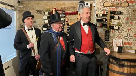 Three men wearing top hats and tail coats. Behind them is a wall with wine on it. To the left is a white door and to the right is a barrel. 