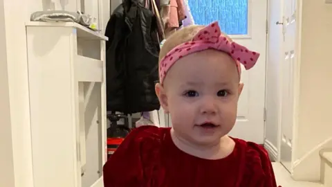 NHS Lanarkshire Aurora El Alti, a baby, in a hallway of a house. She is wearing a red dress and has a pink bow on her head. 