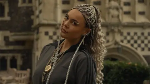 Harriet Laws The photo shows a woman looking sideways at the camera wearing a grey Adidas tracksuit top, and gold necklaces. Her hair is long and braided, and she has a piercing in her eyebrow. She is standing outside and behind her is a hedge and an old building.