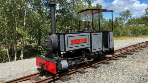 Groudle Glen Railway A small black and grey steam locomotive sits on railway tracks lined with trees on a sunny day. The name Annie is on a red sign on the side, matching a red panel at the front. 
