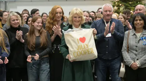 Britain's Queen Camilla holds a tote bag with the name of Jilly Cooper on it, who wrote the original books entitled Rivals, as she poses with members of the cast and crew during a visit to the set of the TV series Rivals.