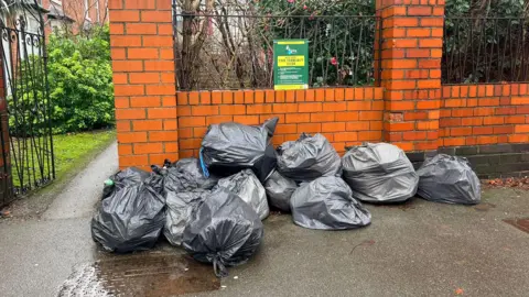 Gabriel Bononi A pile of binbags against an orange brick wall, lying on pavement. There is a gate in the wall which is open, leading to a path