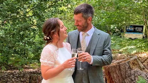 A man, dressed in a suit, and a woman in a wedding dress toast champagne in a woodland.