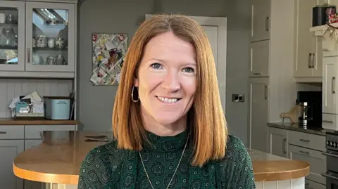 Angela smiling. She has shoulder length light auburn hair. She is wearing a green patterned top and a gold necklace. She is also wearing gold hoop earrings.