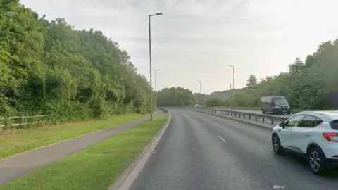 A dual carriageway curves away, with a separate footpath alongside the road. Grass between the road and the footpath. Tall hedges on either side of the road.