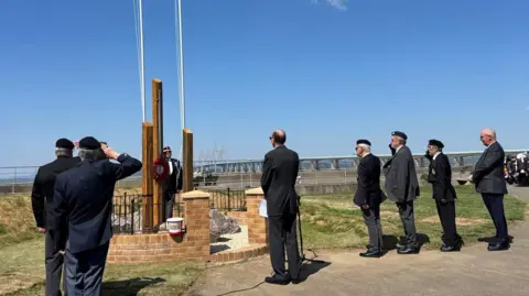 Men in suits and uniform are standing around three pillars that make up a new memorial. They are saluting it.