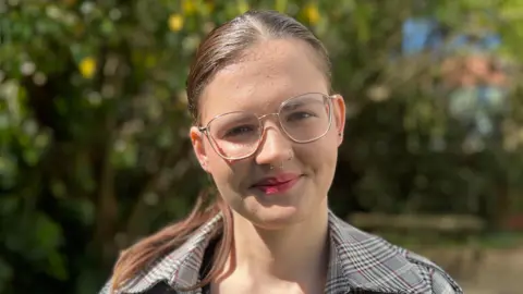 Simon Dedman/BBC Madi Faulkner-Hatt stands outside on a sunny day and smiles directly at the camera. She has dark hair that has been tied back behind her head, she wears glasses and a chequered jacket.