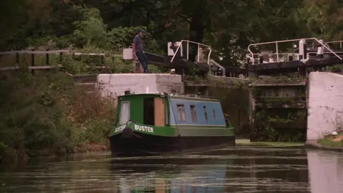 A narrowboat on a canal approaches a lock, while a person stands beside the lock gates. Trees and vegetation line the waterway in the background.