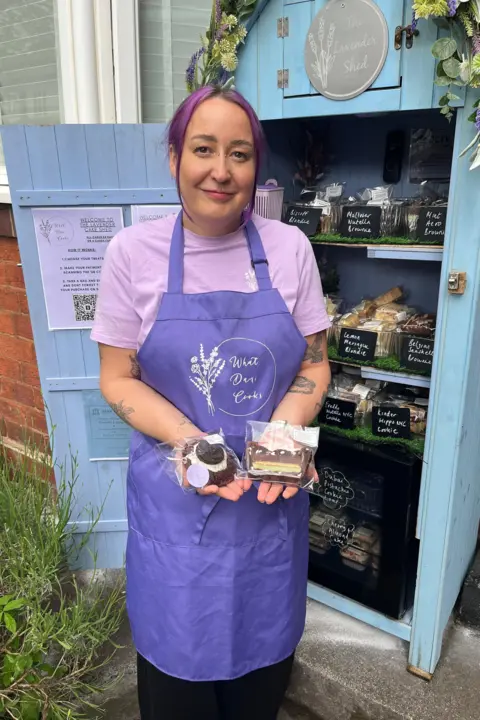 Dani wearing a purple apron and purple t-shirt standing next to the cake shed. The door is open and there are three shelves of cakes. The shed is blue and has a floral garland on the top. She is holding a cake in each hand.