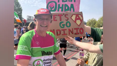 A man during the London Marathon, smiling at the camera. A charity emblem is on his green, pink and white shirt and he is wearing a baseball cap. There are other runners in the background with signs and flags.