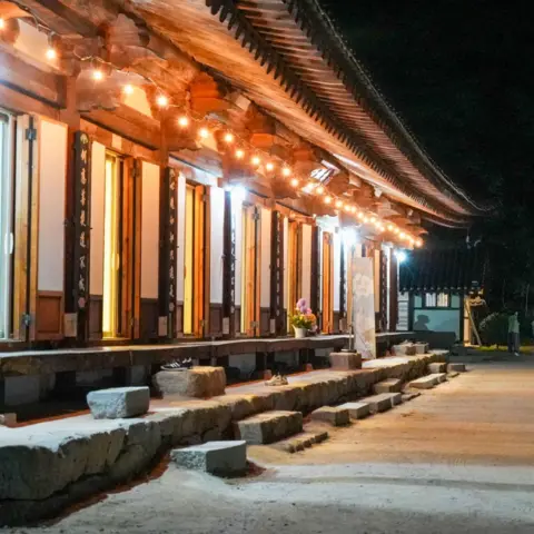 BBC/ Hosu Lee Traditional Korean temple building illuminated by warm lights at night, with a curved tiled roof, wooden columns, and stone steps lining a wide courtyard. Several visitors walk along the path beside the temple.