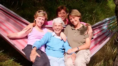 Sally Magnusson Sally Magnusson (left) in brown trousers and a pink T-shirt with sunglasses on her head, with her mother Mamie (centre), wearing a light blue jumper with white blouse underneath, sister Margaret (right) in beige trousers and a brown T-shirt. The three woman are sitting on a red and pink striped hammock with grass around them. Sally's sister Anna is crouching down behind them and is wearing a pink long-sleeved top.