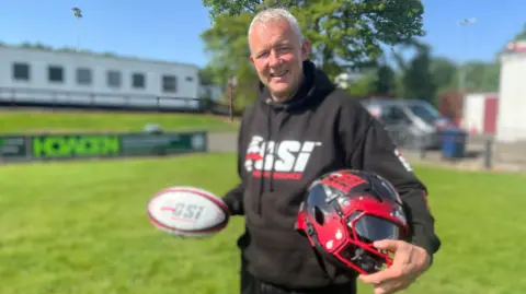 A grey-haired man, wearing a black sweat shirt, holds a rugby ball and American football helmet