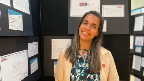 A woman in a cream blazer smiles in front of a children's design display in a school hall.