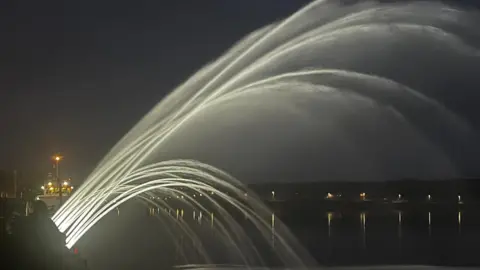 Water shoots out from a quayside fountain at night. The water has been lit up. A boat is in the background.