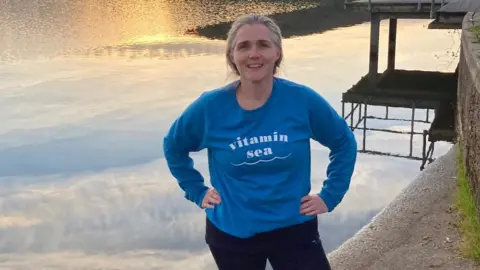 A woman is standing beside a lake with her hands on her hips. She has grey hair that is tied back. She is wearing a long-sleeve blue t-shirt and a pair of black leggings.