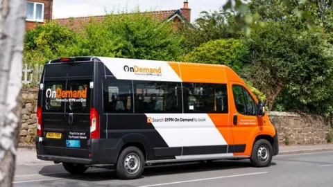 An image of a minibus on a country road. The vehicle is driving away from the picture to the right. It is painted orange black and white with "Search SYPN OnDemand to book" written on the side.