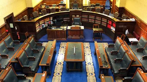 An empty Tynwald chamber, with has blue leather-clad seats on the floor for MHKs and brown leather chairs on an elevated level positioned in a semi-circle behind the Speaker's desk. The Sword of State is on a table in the middle of the room which has a blue and gold carpet.