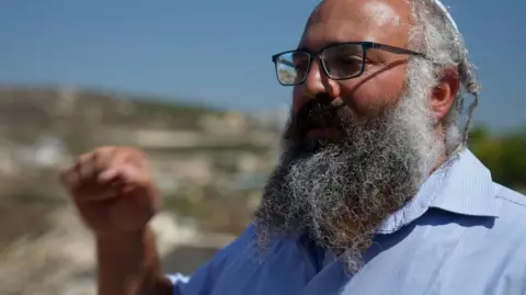 Yehuda Shimon, a large, bearded man wearing glasses, gestures with his hand