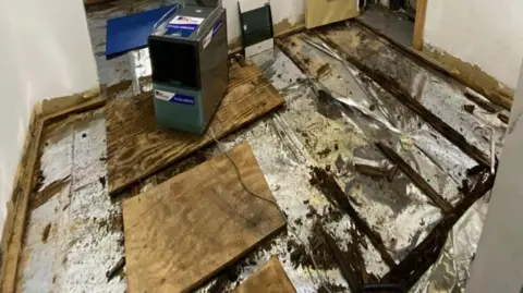 Lincolnshire County Council A classroom interior with the floor covering removed, revealing rotting timber joists, dark mould patches and damp insulation which is silver in colour, with loose wooden boards and a small dehumidifier placed on the exposed, water-damaged floor.