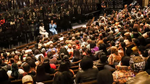 Black British Book Festival Two women are sat in front of a mirrored wall in front of rows of other people sat in seats.