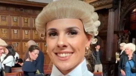 A female barrister in a wig smiles inside what appears to be a grand hall, with crowds of people behind her