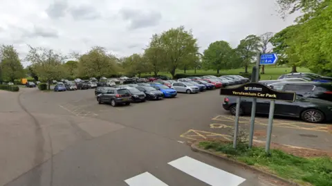 Google Rows of cars parked in a surface car park with trees and an open green space surrounding it.