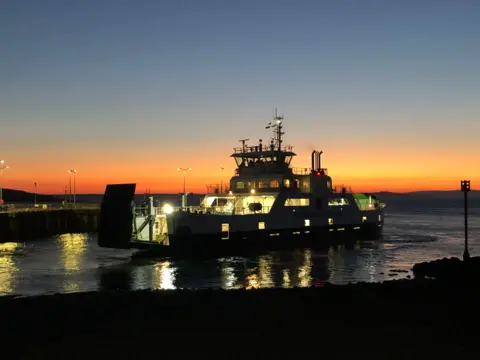 Richard Morris A ferry lit up at night as it sails off, with the sun setting and an orange glow in the sky