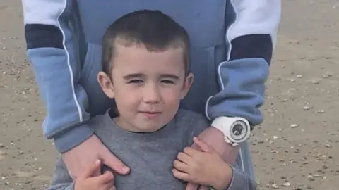 Family handout Lincoln Button, who has short black hair, wearing a grey tracksuit with a slight smile. He is being held from behind by someone, whose face cannot be seen. They have a hoodie on and a large white watch.