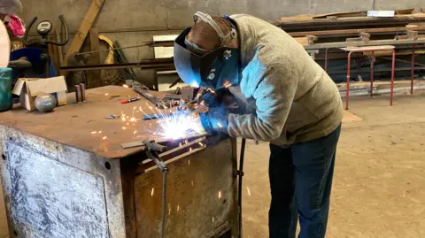 Steven Nesbitt in his workshop welding the metal chains. He is wearing a protective mask and sparks are flying from the welder.