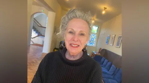 A woman smiles. She has grey hair and is wearing a jumper. 