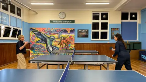 Alexa Rusakoff Former Traitors contestant Amanda Collier and radio presenter Allison Ferns pictured at the Brighton Table Tennis Club. They are playing table tennis. The wall is painted light blue and the canvas says 'Solidarity - Refugees Welcome'. There is a hummingbird and flower painted onto the canvas.