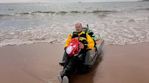 Lee Robinson Lee Robinson sits resting on the back of his jet ski, which is pulled up onto a sandy shoreline with small gentle waves washing around it, holding his red helmet against a yellow waterproof jacket.