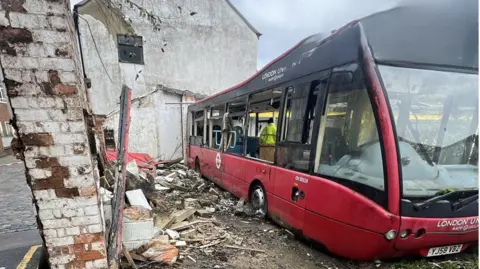 Gloucester City Council The front of the bus as pictured from outside. It is parked slightly diagonally in an abandoned courtyard plot, with a dilapidated brick wall on the left. Down the side of the red bus there is a large pile of rubbish, wood and debris. Several of the windows have been smashed and there is a council worker clearing the inside. 
