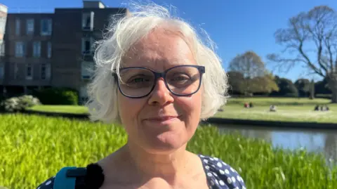 A person with short white hair and glasses standing beside a calm waterway on a sunny day, wearing a blue polka‑dot top with a turquoise bag strap, with a modern building, trees and people relaxing on the grass in the background.