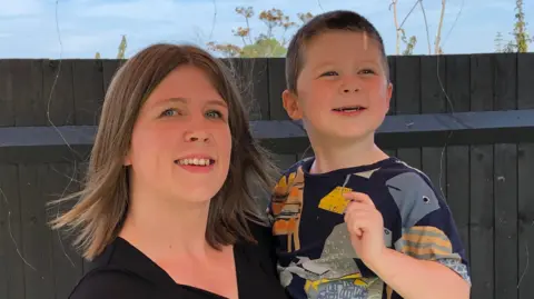 Abbie, in a black top, pictured holding her son Toby, who is wearing a patterned top, in their back garden in front of a wooden fence.