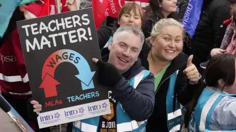 PA Media A man is smiling at the camera while wearing a blue hi-vis vest and holding up a sign saying 'Teachers Matter' along with a graphic showing MLA wages being trickled down to teachers. On his left there is a woman smiling at the camera with her thumb up.