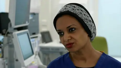 A woman with a black and white head covering and blue scrubs in a clinical setting with hospital equipment visible behind her.