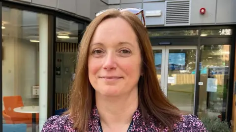A woman with long brown hair wearing a pink and black patterned dress standing outside a building