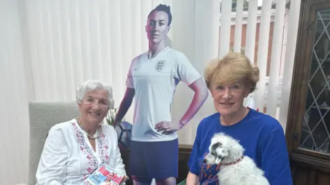 Gloria Stapleton (left) has short white hair and is smiling. She wears a pearl necklace and white cardigan. She is holding a copy of Women's Health magazine, which has a picture of Lucy Bronze on it. Julie Tough (right) has short ginger hair and holds a small white dog and a newspaper. A cardboard cutout of Lucy is behind them.
