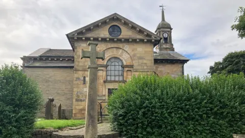 The front of the stone church in Bradford. Three gravestones can be seen next to the entrance and a memorial in front of the stone steps leading up to the entrance. Hedges are on either side. 